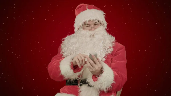 Papai Noel lê e envia mensagens de texto de seu telefone celular no fundo vermelho com neve — Fotografia de Stock