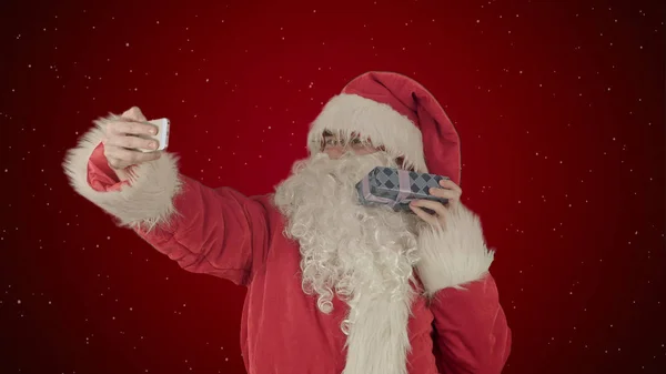 Santa Claus sosteniendo un gran regalo haciendo una selfie en el teléfono inteligente sobre fondo rojo con nieve —  Fotos de Stock