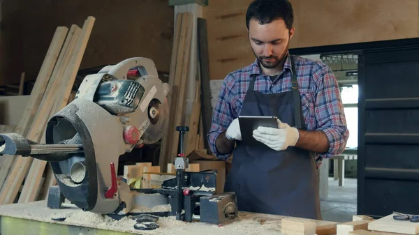 The carpenter works on tablet near electric saw in workshop.