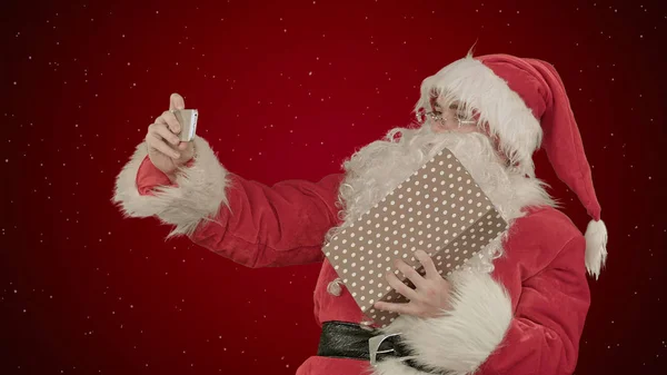 Papai Noel fazer selfie, segurando um grande presente no fundo vermelho com neve — Fotografia de Stock