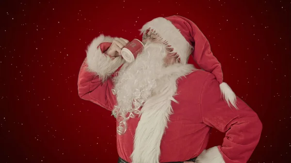 Santa bebe de uma xícara vermelha no fundo vermelho com neve — Fotografia de Stock