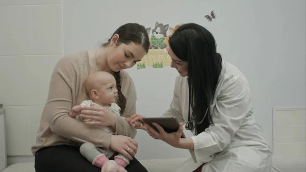 女医は生まれたばかりの赤ちゃんと遊ぶし、母にタブレットを示しています — ストック写真