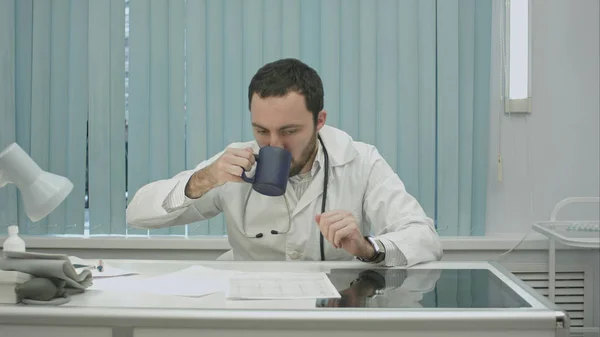 Müde von der Arbeit, bärtiger Arzt trinkt aus Tasse und wacht weiter mit Dokumenten und Röntgenaufnahmen auf — Stockfoto