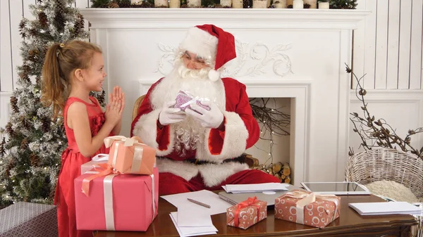 Απασχολημένος Santa ταξινόμησης chidlrens γράμματα ενώ χαριτωμένος μικρός βοηθός του φέρνει ένα δώρο — Φωτογραφία Αρχείου