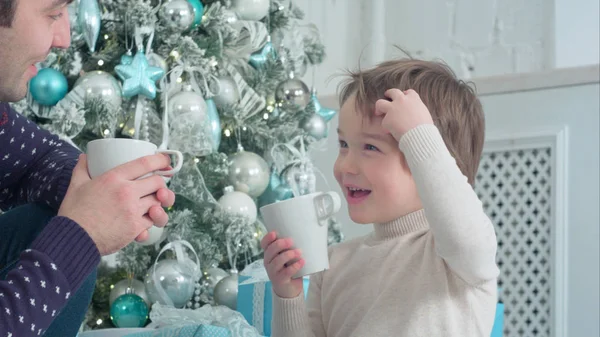 Happy dad and son with cups of tea talking near Christmas tree at home