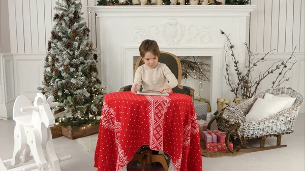 Χαμογελαστό αγόρι παιδί κάθεται στο τραπέζι και να παίζει με το tablet κατά την περίοδο των Χριστουγέννων — Φωτογραφία Αρχείου