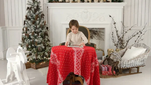 Garoto sorridente sentado à mesa e brincando com tablet durante o Natal — Fotografia de Stock