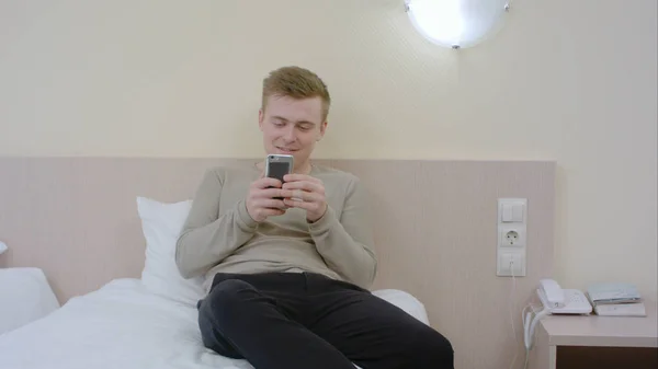Joven guapo usando teléfono inteligente y riendo en el dormitorio en casa — Foto de Stock