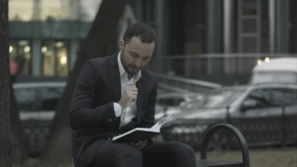 Joven hombre de negocios barbudo pensando y escribiendo notas en Green Park, tráfico detrás — Foto de Stock