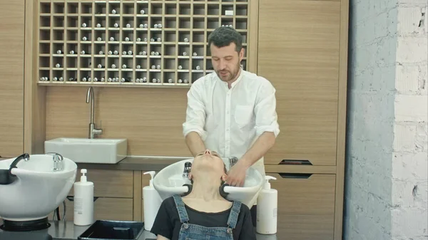 Красивий перукар миє жіноче волосся з концентрацією. Він тримає водопровідний кран. Жінка спирається головою на раковину . — стокове фото