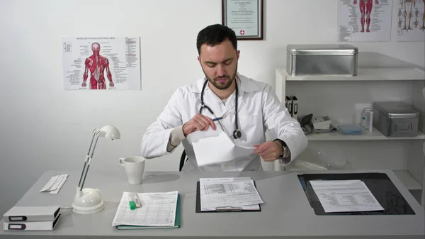 Giovane medico serio distrugge i documenti seduti nello studio medico con cartella — Foto Stock