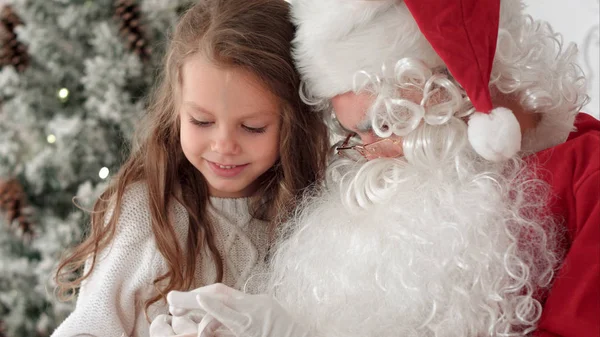 Santa Claus δένοντας μια κορδέλα τόξο για ένα δώρο για ένα μικρό κορίτσι που κάθεται κοντά το χριστουγεννιάτικο δέντρο στο σπίτι — Φωτογραφία Αρχείου