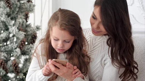 Молодая мать учит свою дочь, как использовать смартфон в комнате, украшенной под Рождество — стоковое фото
