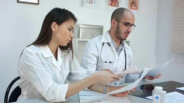 Dos médicos ocupados trabajando con papeles e imágenes de rayos X, respondiendo llamadas telefónicas, recetando pastillas en su escritorio — Foto de Stock