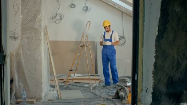 Молодий чоловік у шоломі інженера з безпеки жовтого кольору з використанням мобільного телефону або смс на будівельному майданчику — стокове відео