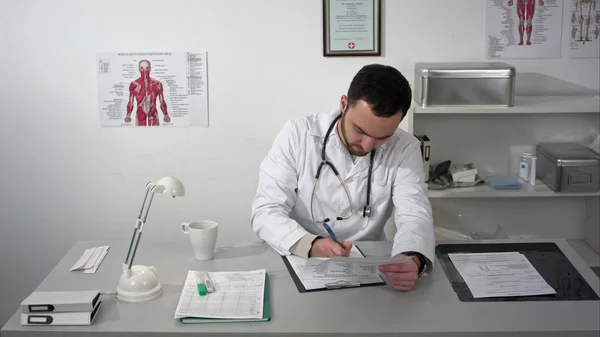 Jovem médico sério sentado em consultório médico com pasta e documentos de leitura — Fotografia de Stock