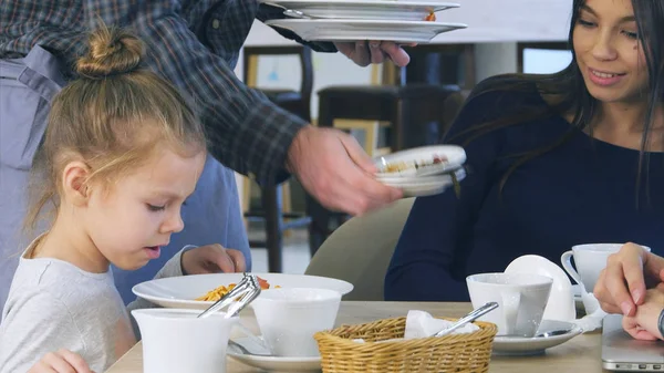 Cute little girl pouring cheese in her spaghetti in italian restaurant while her young parents have ate their vegetable salads yet.