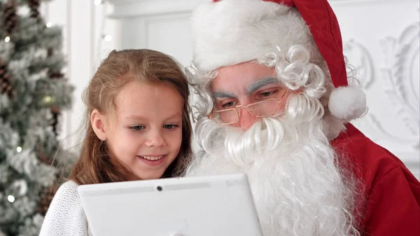 Happy little girl showing surprised Santa presents on the tablet