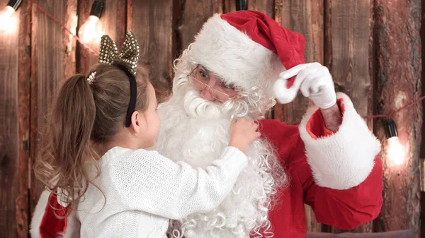 Cute girl playing with Santas beard sitting on his lap