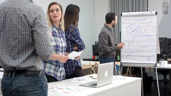 Empresário sorridente apontando para diagramas em flip board enquanto seus colegas oferecendo ideias — Fotografia de Stock