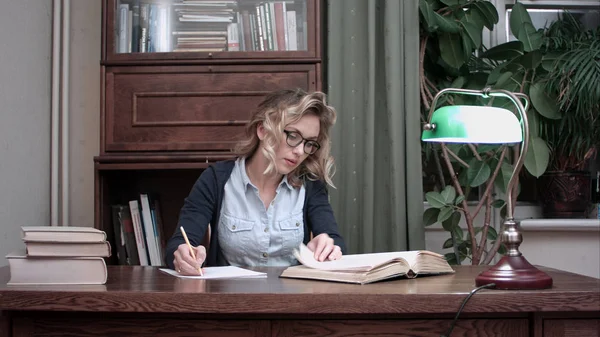 Jovem estudante de óculos sentado em uma mesa e escrevendo notas de um livro — Fotografia de Stock