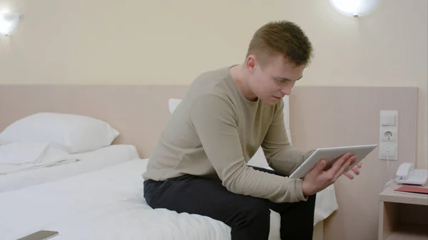 Молодой человек с помощью современного планшета сидит на кровати в гостиничном номере — стоковое фото