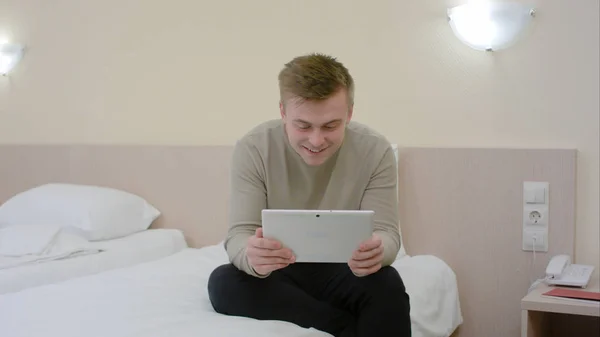 젊은 남자가 그냥 호텔 방에 오고, 침대에 앉아서 태블릿에 화상 채팅을 하는 — 스톡 사진