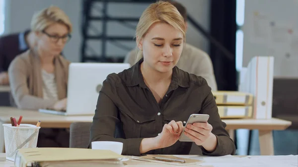 Allegro giovane bella donna utilizzando il suo smartphone con il sorriso mentre seduto al suo posto di lavoro — Foto Stock