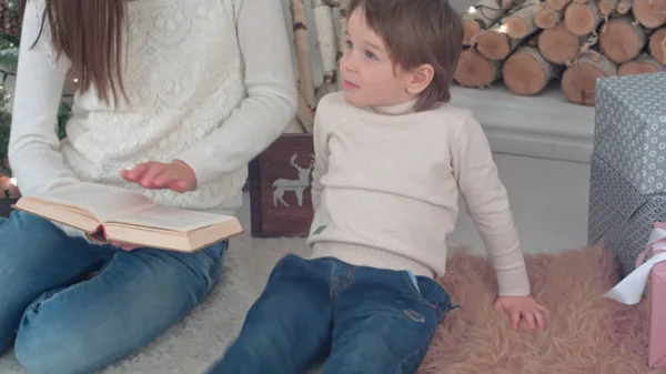 Caprichoso niño escuchando a su mamá leyendo un libro cerca del árbol de Navidad — Foto de Stock