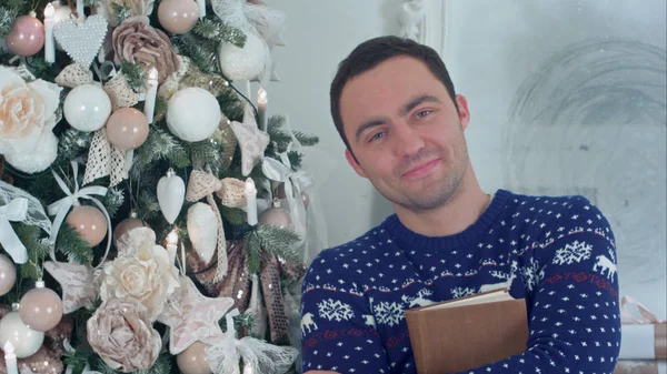 Joven sonriente sosteniendo un libro de pie junto a un árbol de Navidad — Foto de Stock