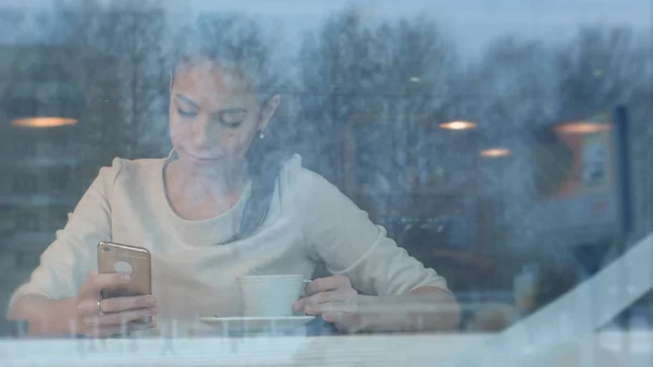 Mujer bonita sonriente con una taza de té mensajes de texto en el teléfono inteligente en un café — Foto de Stock