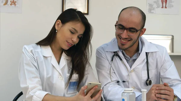 Ganska ung sjuksköterska visar något roligt på sin telefon till manliga kollega — Stockfoto