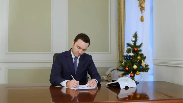 Hombre de negocios leyendo y firmando documentos sentados junto a la mesa en la oficina cerca del árbol de Año Nuevo —  Fotos de Stock