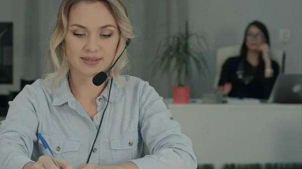 Consultora sonriente con auriculares haciendo notas mientras su compañera de trabajo se toma selfies — Foto de Stock