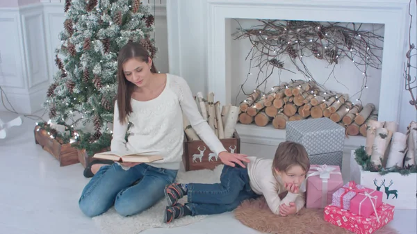 Mother reading a book for her little son lying near Christmas tree