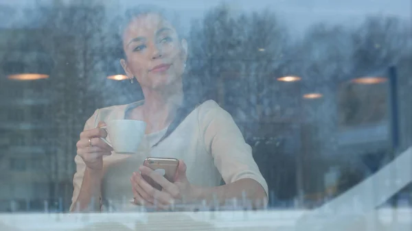 Menina bonita usando telefone enquanto bebe café em um café visto através da janela — Fotografia de Stock