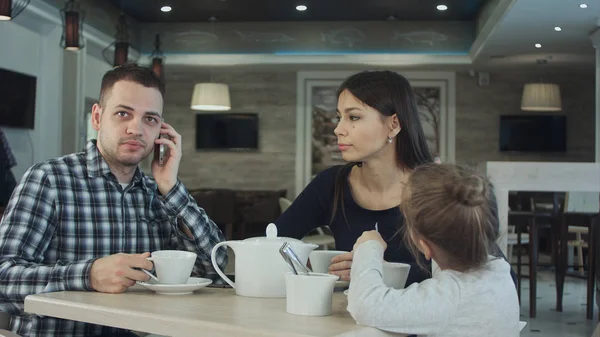 Ung familj i café. Far ringa ett telefonsamtal medan mor och dotter talar. Mamma tittar på honom missnöjd. — Stockfoto