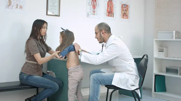 Manlig barnläkare med stetoskop lyssnar på lilla flickan tillbaka medan mor håller hennes hår — Stockfoto