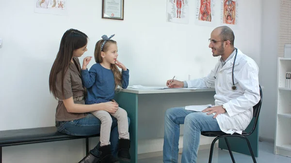 Un médecin souriant parle avec une petite fille timide assise sur ses tours de maman — Photo