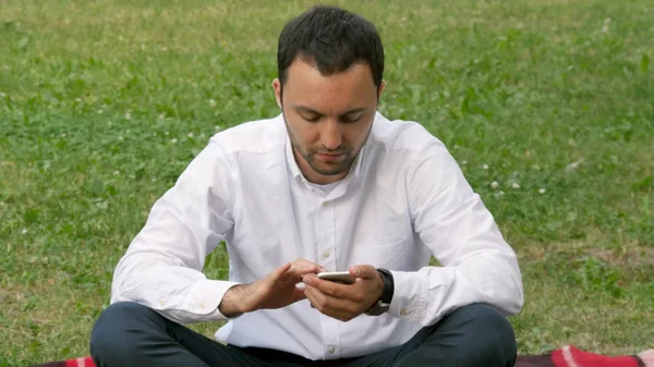 Красивый взрослый мужчина отправляет смс с мобильного телефона в солнечный вечер на открытом воздухе в парке — стоковое фото