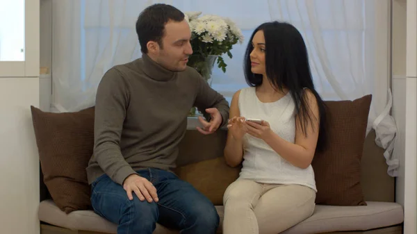 Young man and woman sitting on sofa in living room and discussing something on screen of smart phone