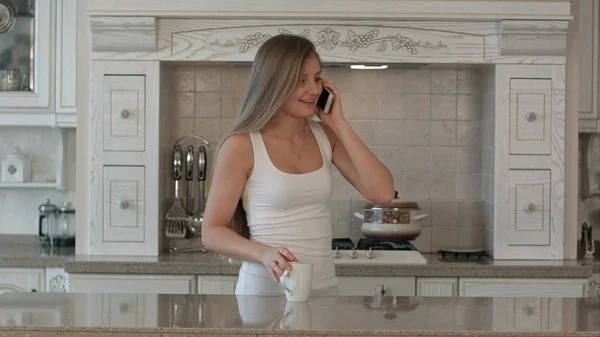 Молода красива жінка говорить мобільний телефон, п'є каву або чай на кухні, щаслива посмішка — стокове фото