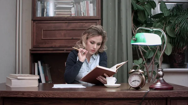 Giovane donna concentrata che legge un libro con una matita in mano, seduta a una scrivania quando il suo collega maschio le porta un altro mucchio di libri — Foto Stock
