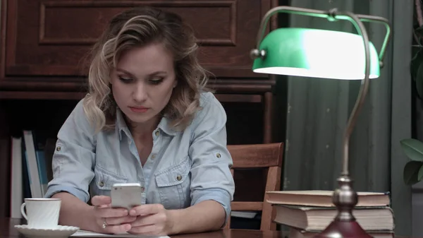 Серйозна молода жінка сидить на робочому місці і друкує повідомлення на своєму телефоні — стокове фото