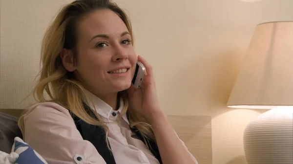 Молодая женщина имеет положительный разговор с помощью мобильного телефона сидя на кровати — стоковое фото