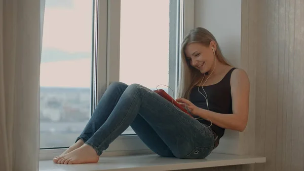 Menina senta-se na janela ouvir música e trabalha com tablet — Fotografia de Stock