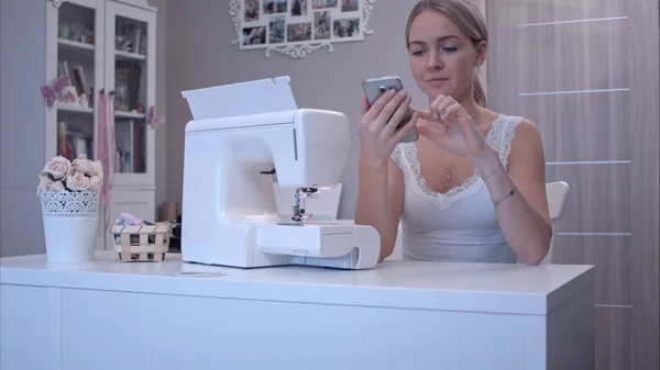 Mujer joven con teléfono sentado en la mesa al lado de la máquina de coser — Foto de Stock