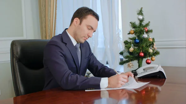 Jovem empresário lendo documentos e assinando perto da mesa da Árvore de Ano Novo — Fotografia de Stock