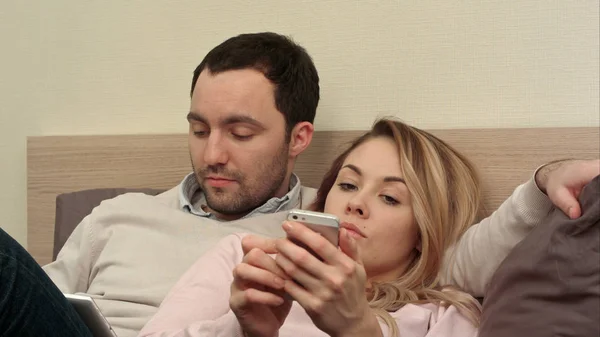 Молодая пара лежит в постели, мужчина с цифровым планшетом, скучающая женщина с помощью смартфона — стоковое фото