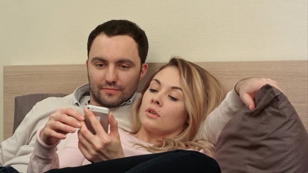 Hermosa pareja sentada en la cama y mirando la foto del viaje en el teléfono inteligente, tocando la pantalla — Foto de Stock